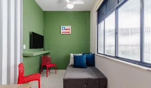A room with a green wall, a ceiling fan, a TV on a shelf, a window with blinds, a sofa with blue cushions, and two red chairs. - Studio the City Center 300m Pelourinho Hai0203 (Salvador)