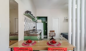 A compact kitchen with a refrigerator, stove, and shelves. A dining table with red chairs and a small table lamp is in the foreground. - Studio the City Center 300m Pelourinho Hai0203 (Salvador)