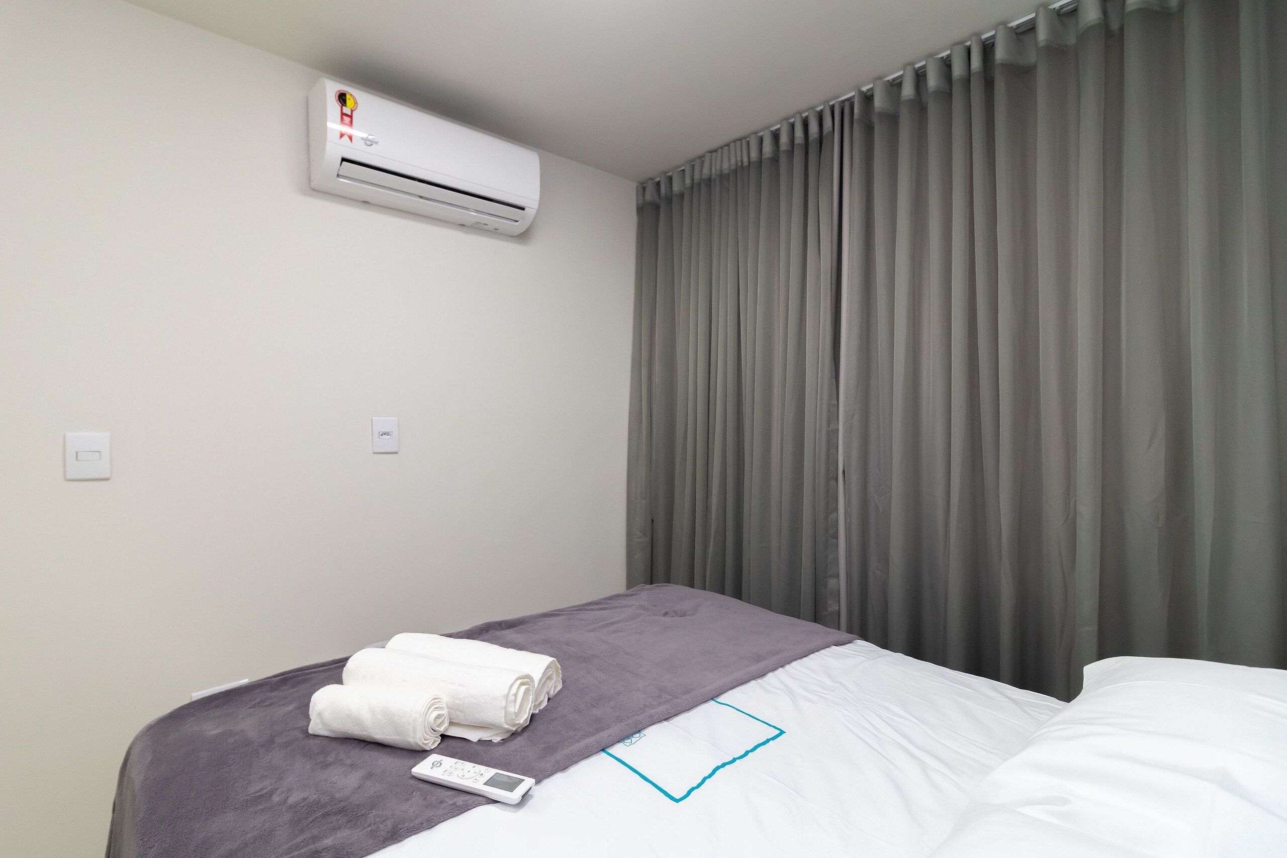 Ein Hotelzimmer mit einem Bett, einer grauen Decke, einem aufgerollten weißen Handtuch, einer Klimaanlage und grauen Vorhängen.