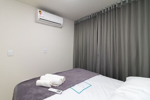 Ein Hotelzimmer mit einem Bett, einer grauen Decke, einem aufgerollten weißen Handtuch, einer Klimaanlage und grauen Vorhängen.