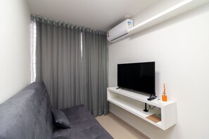 A modern living room with a grey sofa, a flat-screen TV on a white shelf, and a window with grey curtains. - Apart in Asa Norte, 750m From the Mall Nor0201 (Brasília)