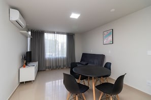 A modern living room with a round table, black chairs, a white sideboard, a flat-screen TV, and a large window with grey curtains. - 1BR Apartment, Self-checkin, Asa Norte Nor0202 (Brasília)