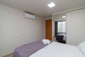 Habitación de hotel con cama, colcha morada, pared blanca, unidad de aire acondicionado, puerta y espejo que refleja una ventana con cortinas.