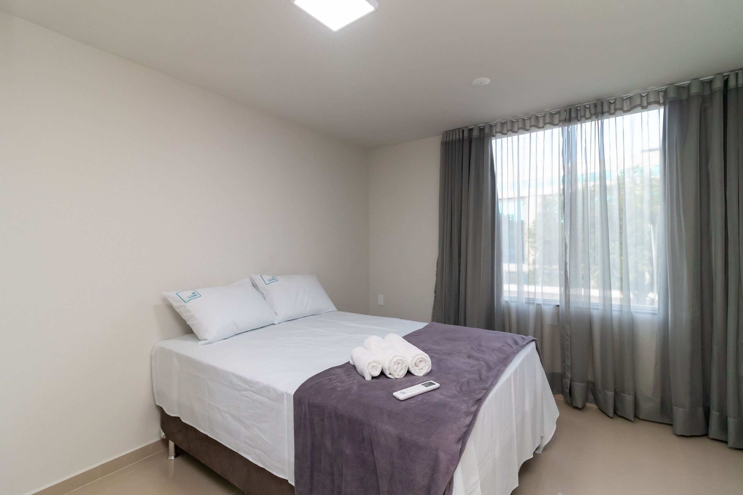 Habitació d'hotel amb un llit, roba blanca, una manta morada, tovalloles arremolinades, un comandament a distància i una finestra amb cortines grises.