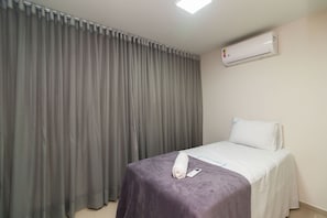 Hotellihuone, jossa on sänky, rullalle taitettu pyyhe, kaukosäädin ja ilmastointilaite seinässä.