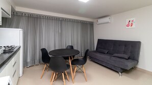 Un espacio habitable compacto con kitchenette, una zona de comedor con una mesa redonda y cuatro sillas, un sofá gris oscuro y una cortina gris que cubre la ventana.