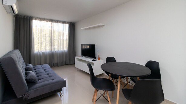 Ein modernes Wohnzimmer mit einer Couch, einem Fernseher auf einem weißen Schrank und einem runden Tisch mit Stühlen.