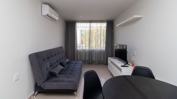 A modern, minimalistic room with a grey sofa, a TV, a small table, and a window with curtains. - 1BR Fully Equipped Apartment, Asa Norte Nor0204 (Brasília)