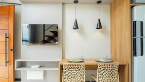 A compact kitchen with a wooden dining table, two pendant lights, a built-in TV, and a refrigerator.