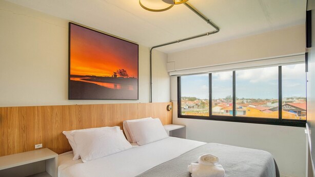 A hotel room with a bed, a nightstand, a wall-mounted painting, a window with a view, and a ceiling light fixture.