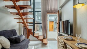 Una sala de estar moderna con una escalera de madera, un sofá, un comedor con televisor y vistas por una ventana grande.