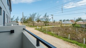 Un balcón con barandilla de metal que da a un sendero de tierra, vegetación escasa y edificios a lo lejos.