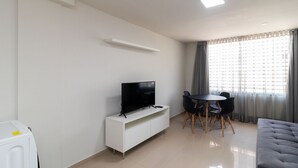 A modern living room with a flat-screen TV on a white console, a dining area with a round table and chairs, and a large window with curtains. - 1BR Apartment, Self-check-in, Asa Norte Nor0203 (Brasília)