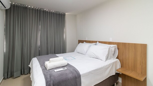 A neatly made bed with a remote control, a wooden nightstand, and a grey curtain. - 1BR Apartment, Self-check-in, Asa Norte Nor0203 (Brasília)