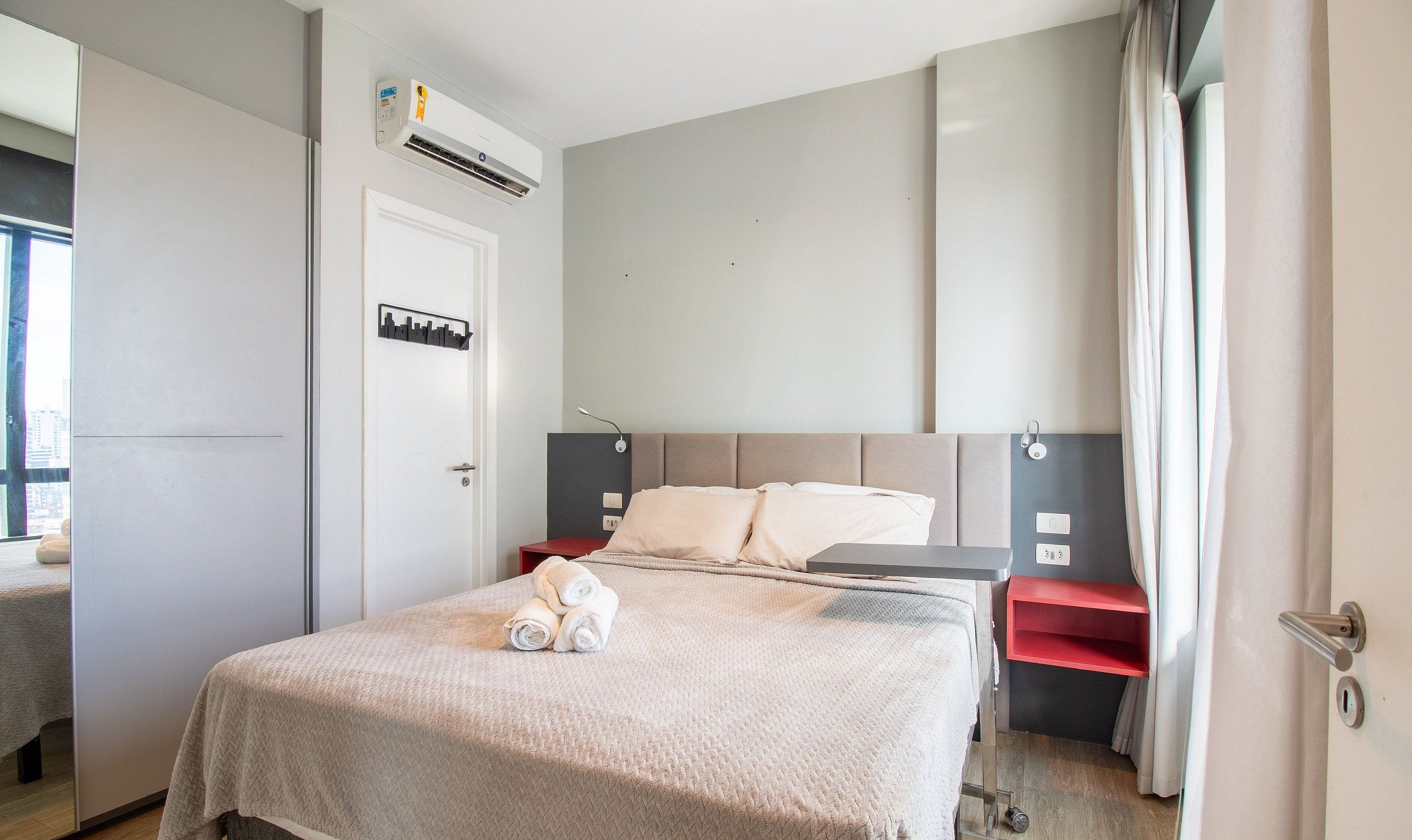 Ein Hotelzimmer mit Bett, Nachttisch, rotem Beistelltisch, Wand-Klimaanlage und einer Tür mit Stadtblick-Hinweis.