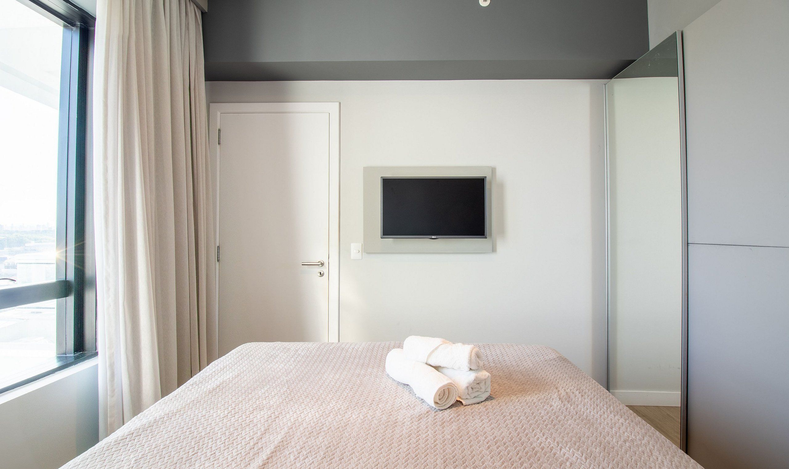Une chambre à coucher avec un lit, un téléviseur fixé au mur et une fenêtre avec des rideaux.