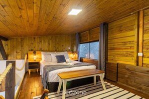 Una habitación de cabaña de madera con cama, mesitas de noche, un banco y una ventana.