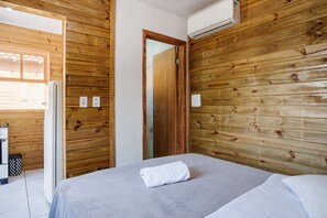 A room with wooden walls, a bed with white bedding, and an air conditioning unit mounted on the wall. - Cozy Cabin 10 min From Praia do Rosa Prj025 (Imbituba)