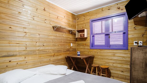 A wooden room with a bed, a small table, a stool, and a window with shutters. - Cozy Cabin 10 min From Praia do Rosa Prj025 (Imbituba)