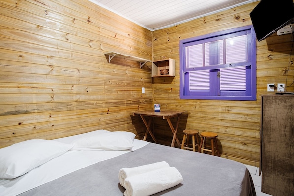 A wooden room with a bed, a small table, a stool, and a window with shutters. - Cozy Cabin 10 min From Praia do Rosa Prj025 (Imbituba)