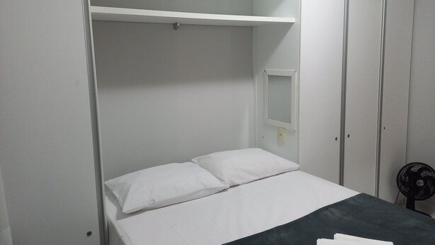 Un letto singolo con biancheria bianca, una coperta scura e un comodino con una ventola.
