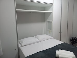 En säng med plats för en person, med vita sängkläder, ett mörkt överkast och ett litet nattduksbord med en fläkt.