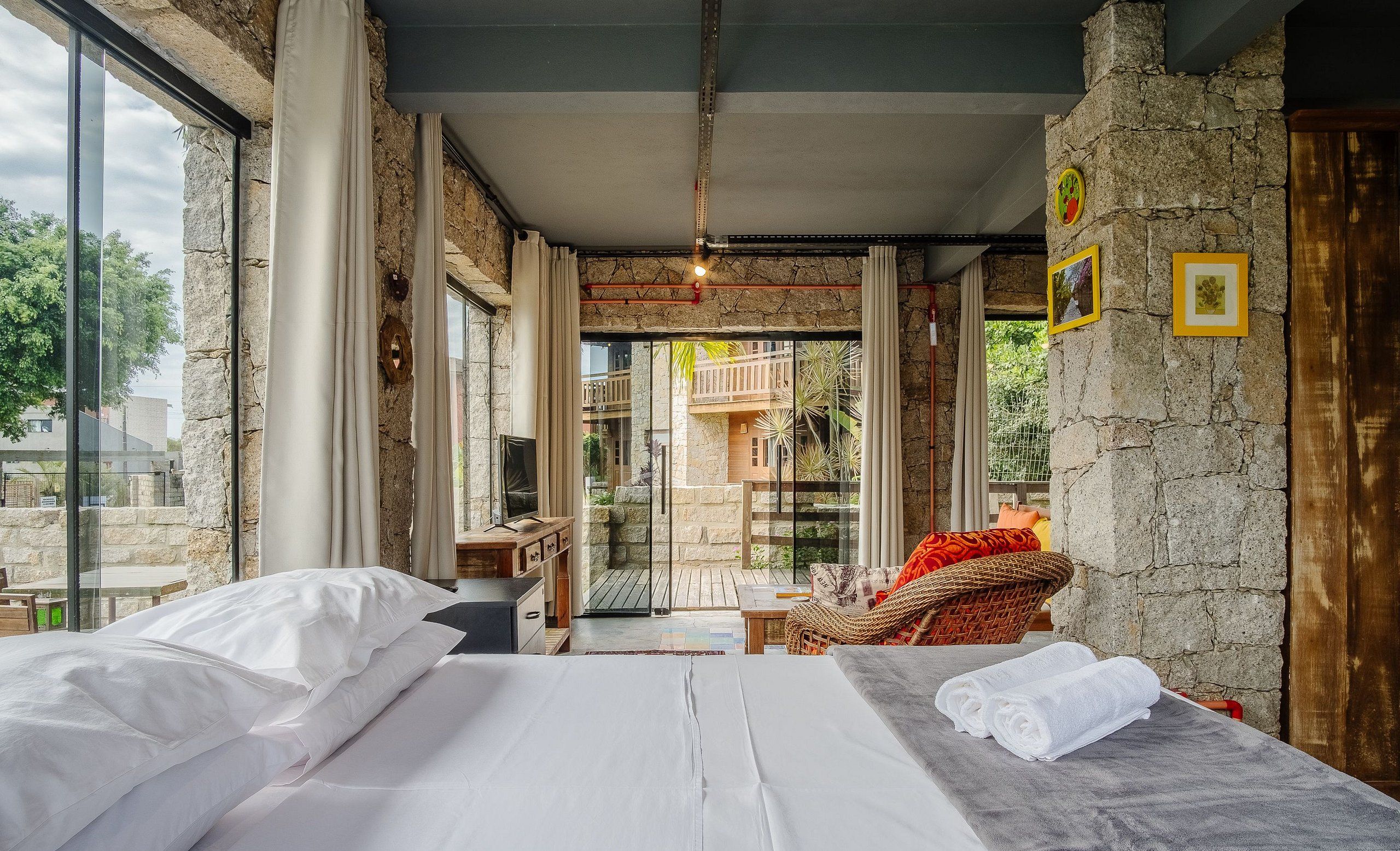 Quarto com cama, uma mesinha e uma cadeira. Tem uma janela com cortinas e uma parede de pedra com quadros.