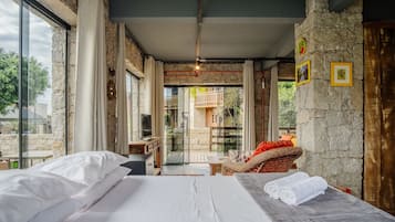 Quarto com cama, uma mesinha e uma cadeira. Tem uma janela com cortinas e uma parede de pedra com quadros.