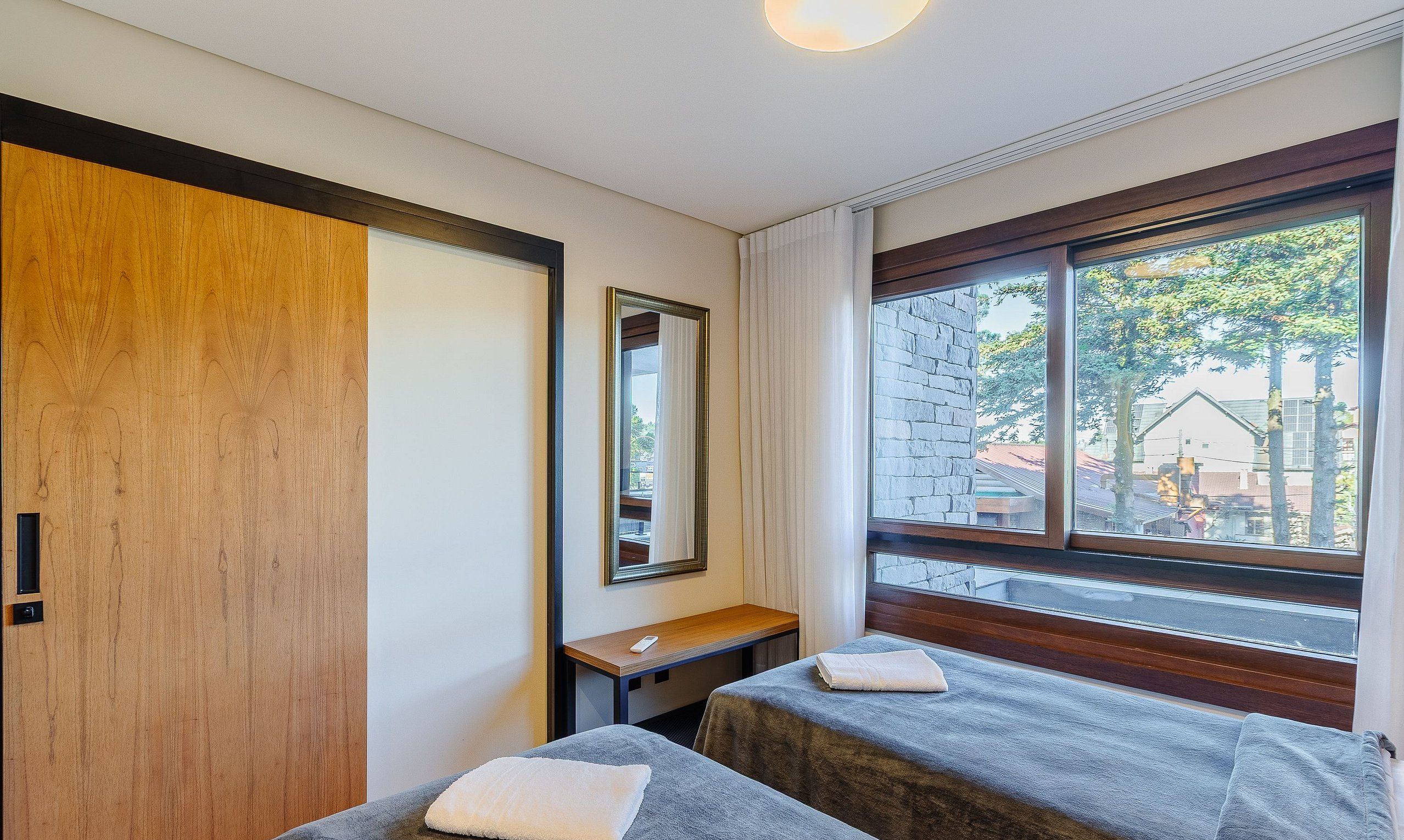 A room with two beds, a wooden door, a mirror, a small table, and a window with a view of buildings and trees.