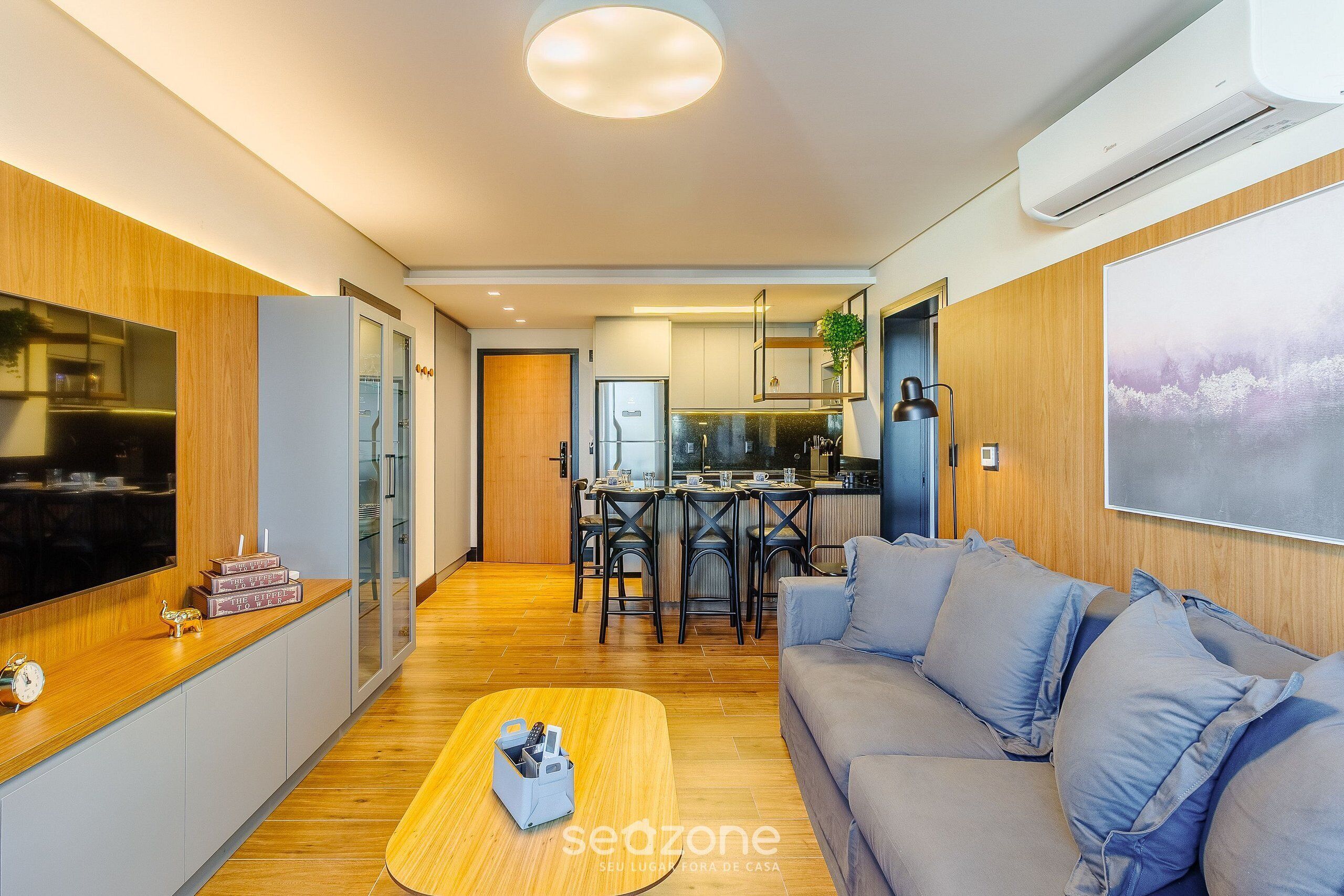 Uma sala de estar moderna com um sofá, uma mesa de centro e uma televisão. Há uma área de cozinha com uma mesa de jantar e cadeiras.