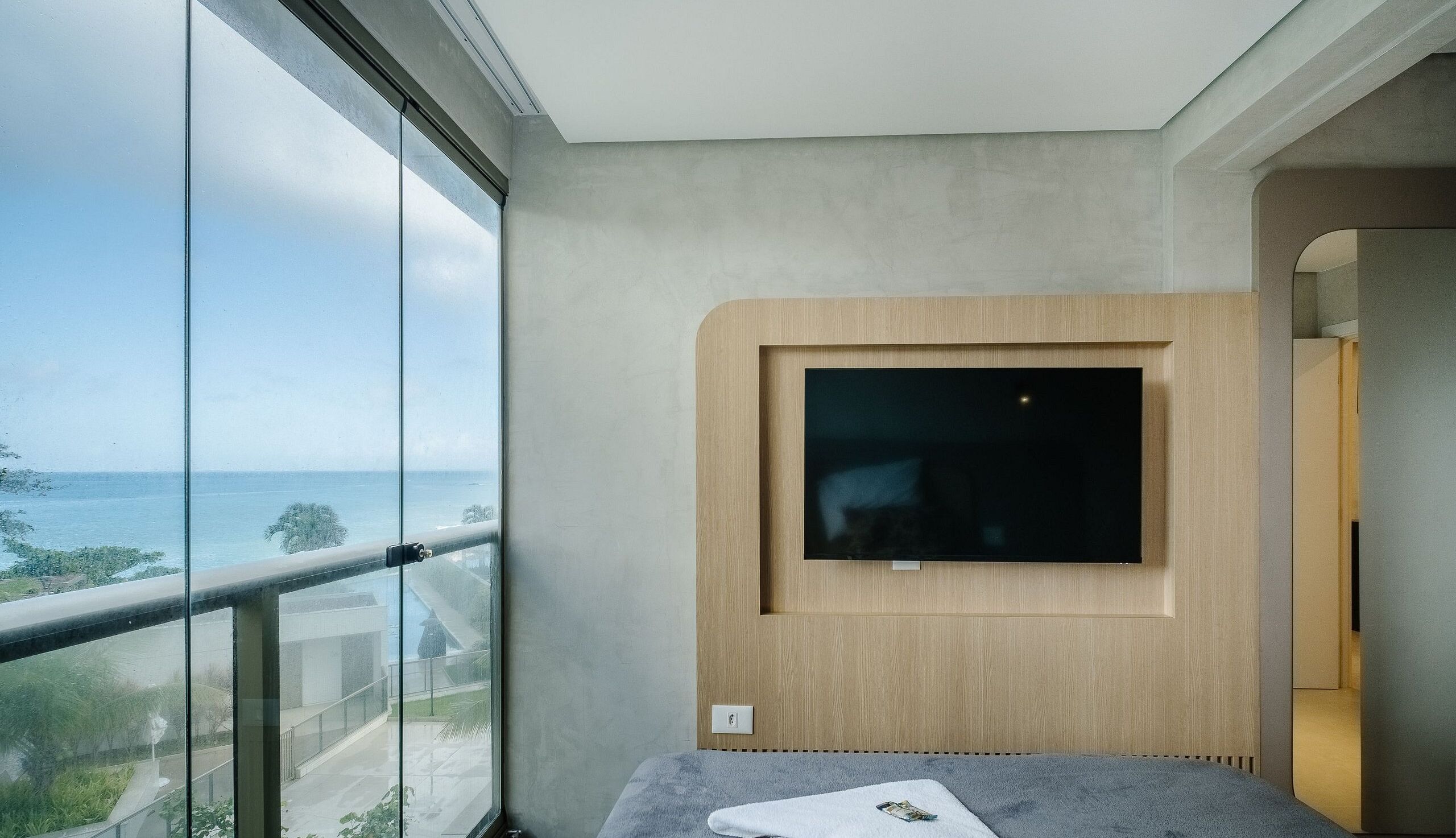 A modern hotel room with a large window offering a view of the sea, a flat-screen TV mounted on a wooden panel, and a small bedside table with a book and remote control.