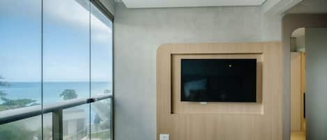 A modern hotel room with a large window offering a view of the sea, a flat-screen TV mounted on a wooden panel, and a small bedside table with a book and remote control.