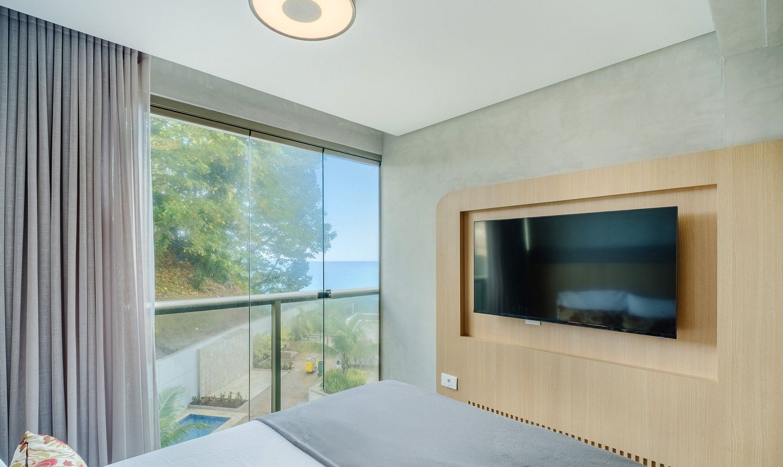 A modern hotel room with a large TV mounted on the wall, a balcony with a view of greenery, and a large window with curtains.