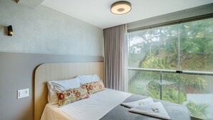 Ett modernt hotellrum med en stor säng, utsikt över grönområden genom stora fönster som sträcker sig från golv till tak, och en taklampa.