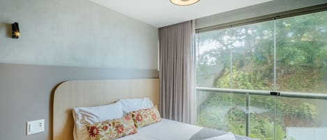 A modern hotel room with a large bed, a view of greenery through floor-to-ceiling windows, and a ceiling light.