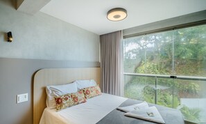 Ein modernes Hotelzimmer mit einem großen Bett, Blick ins Grüne durch raumhohe Fenster und einer Deckenleuchte.
