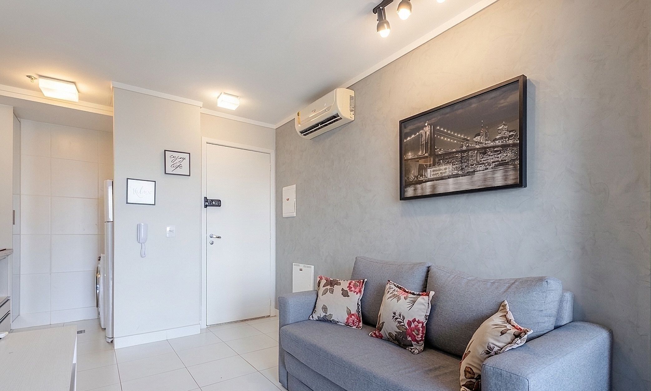 A modern living room with a grey sofa, floral cushions, a framed cityscape picture, and an air conditioning unit.