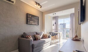 Ein modernes Wohnzimmer mit grauem Sofa, einem Esstisch mit Stühlen, einem großen Fenster mit Vorhängen und einem an der Wand befestigten Fernseher.