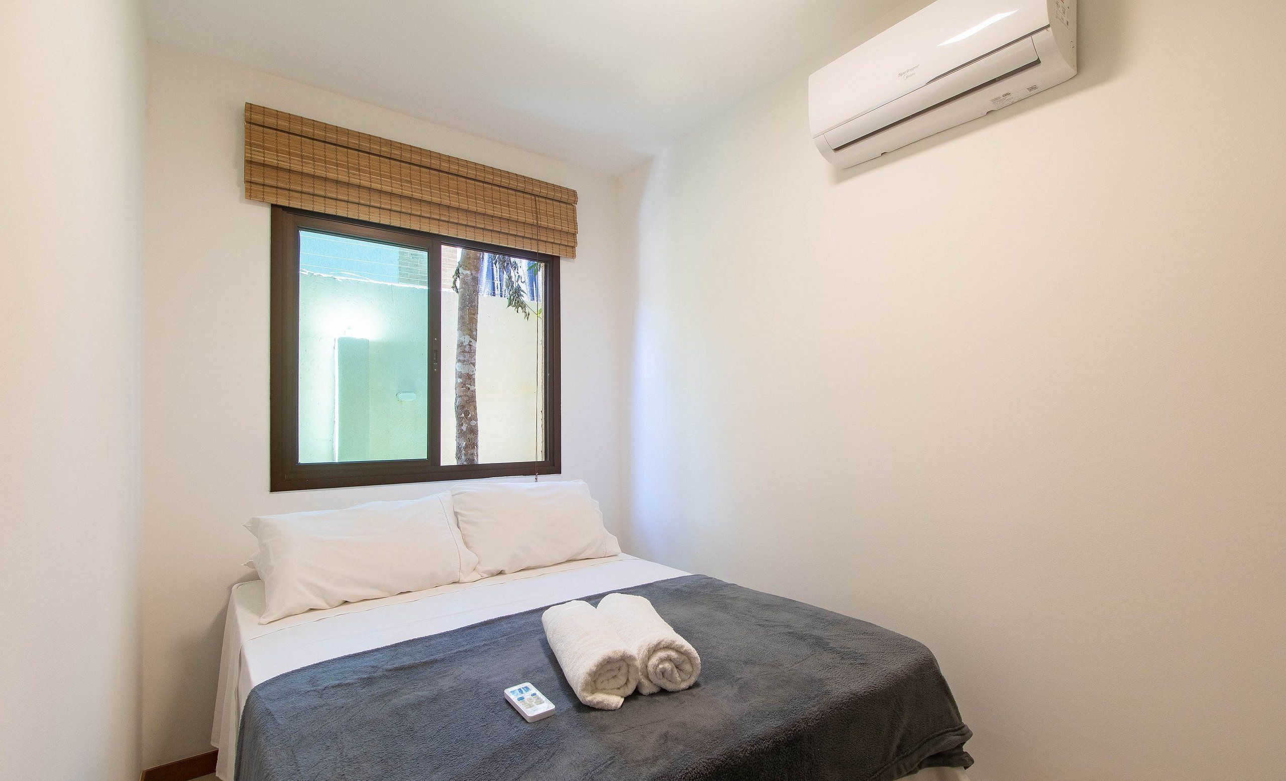 A small, clean room with a bed, a window with blinds, and an air conditioning unit on the wall.