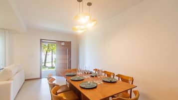 A dining area with a wooden table set for six, surrounded by six wooden chairs. There is a door leading outside, and a white sofa is visible in the background.