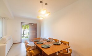 A dining area with a wooden table set for six, surrounded by six wooden chairs. There is a door leading outside, and a white sofa is visible in the background.