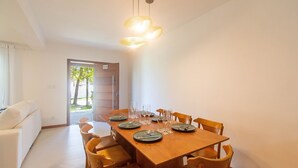Uma sala de jantar com uma mesa de madeira posta para seis pessoas, rodeada por seis cadeiras de madeira. Há uma porta que dá para o exterior, e um sofá branco é visível ao fundo.
