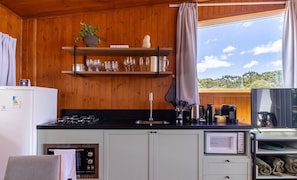 A compact kitchen with wooden walls, a built-in microwave, a gas stove, and a window with a view of trees. - Charming Vale Cabin in Urubici-sc Par0001 (Urubici)