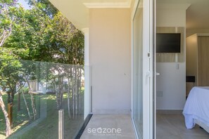 A balcony with a glass door, a bed with a white bedspread, and a mounted TV. - House With Pool in Arraial D'ajuda Mgi024 (Porto Seguro)
