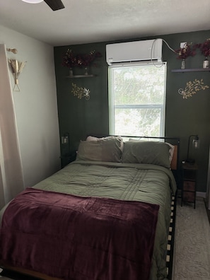A bedroom with a bed, a window with blinds, a bedside lamp, and a wall-mounted air conditioner. - Perfect 2-bedroom cottage with AC in peaceful Livingston (Livingston)