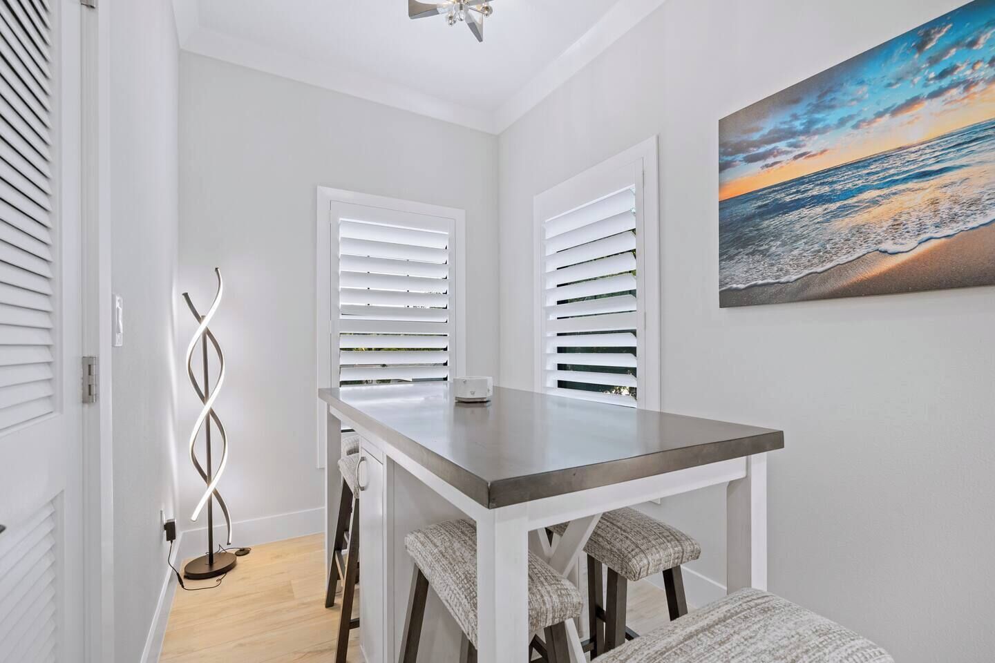 A modern dining area with a table and chairs, white walls, and a painting of a sunset over the ocean.