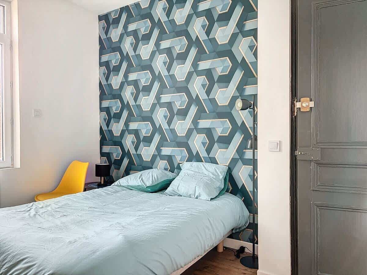 A bedroom with a geometric patterned wall, a bed with light-colored bedding, a yellow chair, and a closed door.