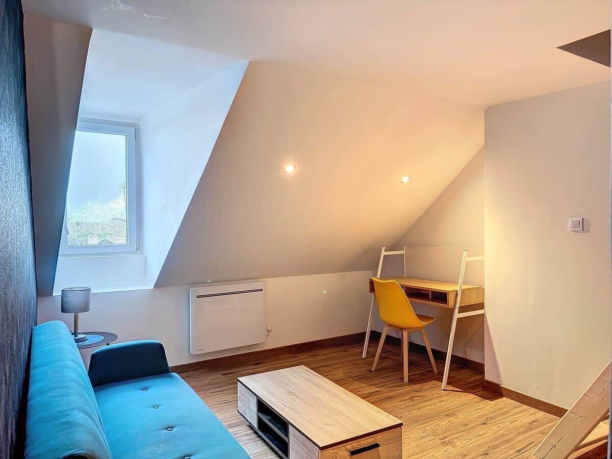 Une pièce de style « loft » avec un plafond en pente, un canapé bleu, un bureau en bois, une chaise jaune et une petite table.