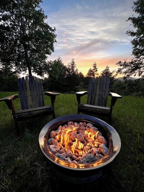 Un foyer avec des bûches qui brûlent, entouré de deux fauteuils Adirondack, dans un cadre extérieur verdoyant.