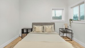 A bedroom with a bed, two bedside tables, a window with blinds, and a small plant on the window sill.